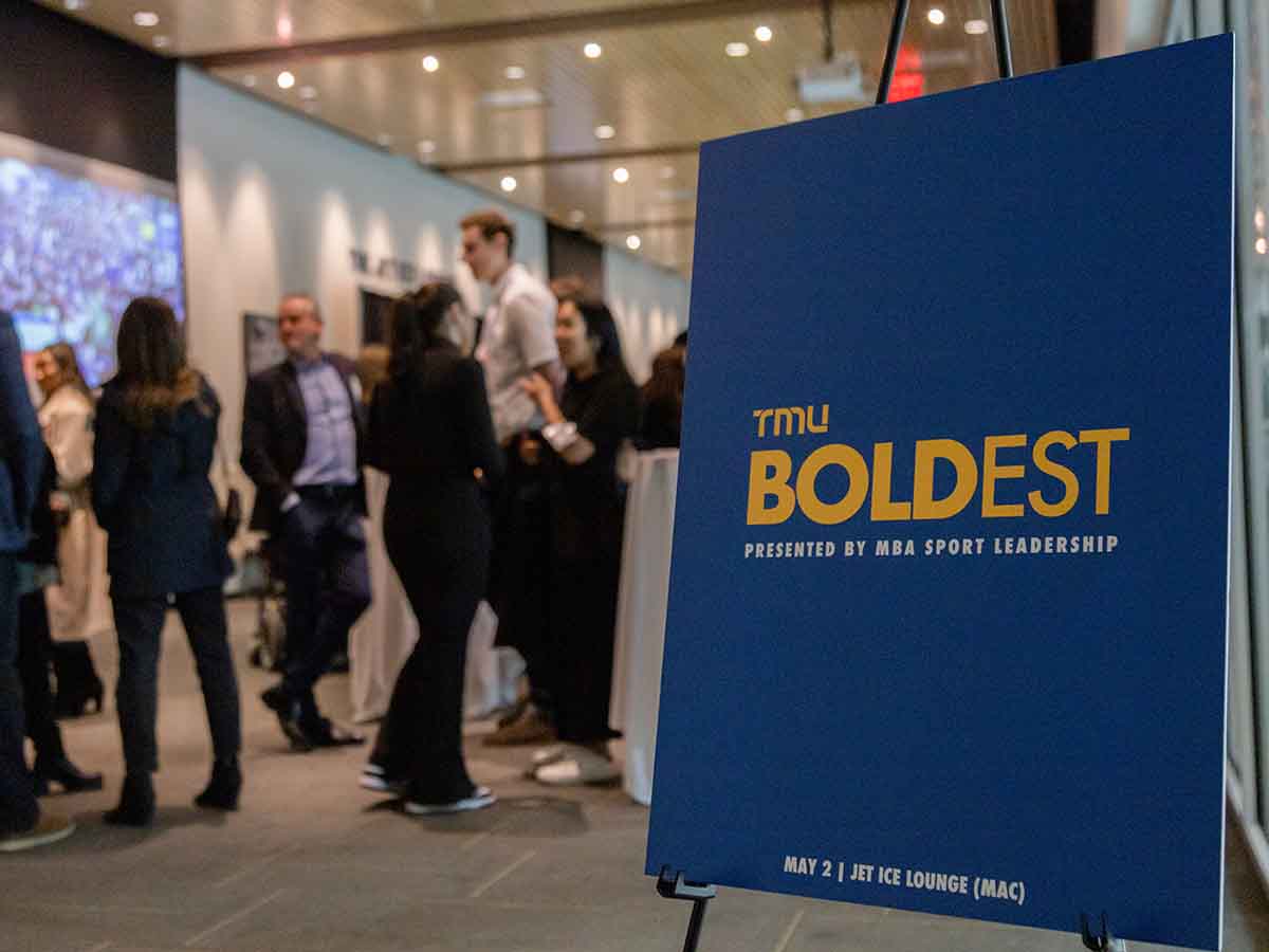 Close up of TMU Boldest Presented by MBA Sport Leadership sign with crowd standing in the background
